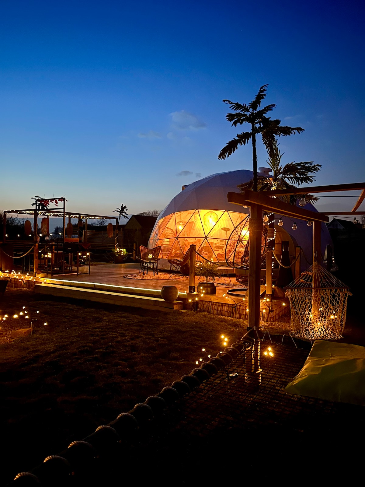 A striking glamping dome is illuminated as dusk falls, surrounded by soft lighting. The structure features a transparent design, allowing glimpses of the interior. Nearby, a comfortable hammock and outdoor seating enhance the relaxing outdoor atmosphere, complemented by palm trees in the background.