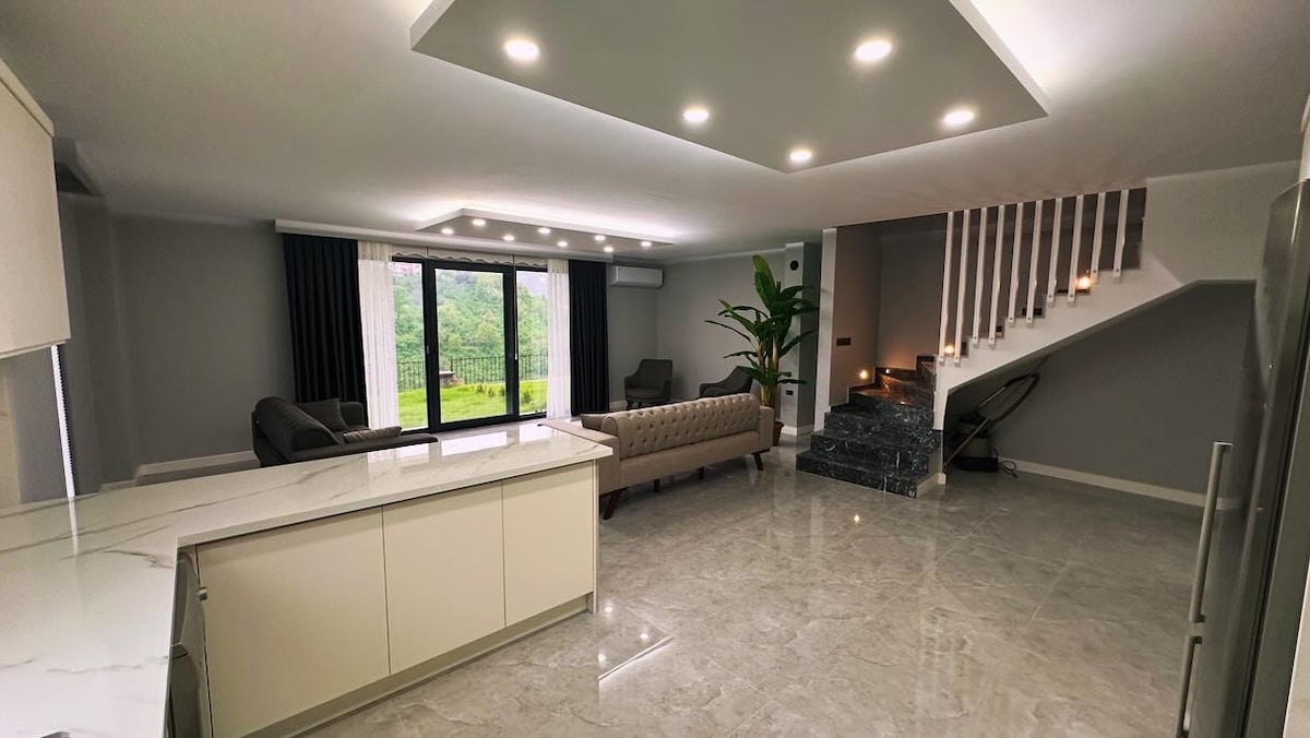 A spacious living area is showcased with natural light flooding through large glass doors, leading out to a green outdoor space. Modern furniture, including a sleek couch and armchairs, contributes to the open layout, while a staircase is visible in the background.