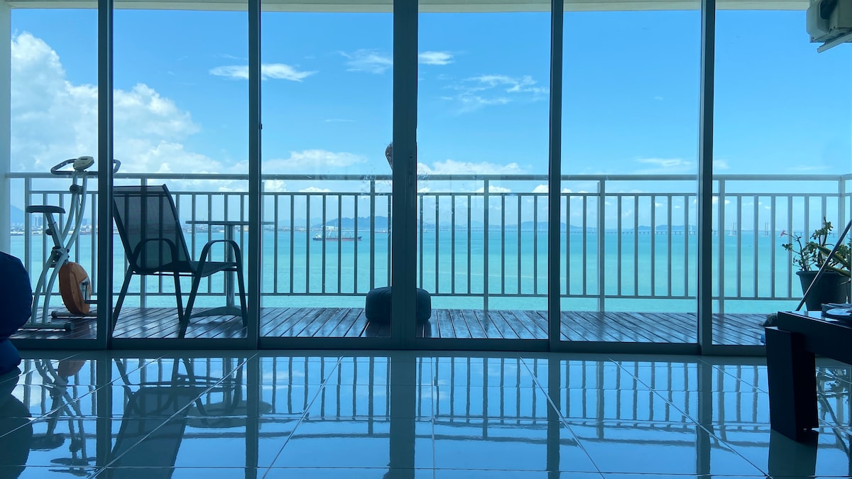 Floor-to-ceiling glass doors offer a panoramic view of the ocean. A reflection of the azure water is visible on the polished tiles. A minimalistic outdoor seating area and exercise bike are positioned on the balcony, complementing the coastal scenery.