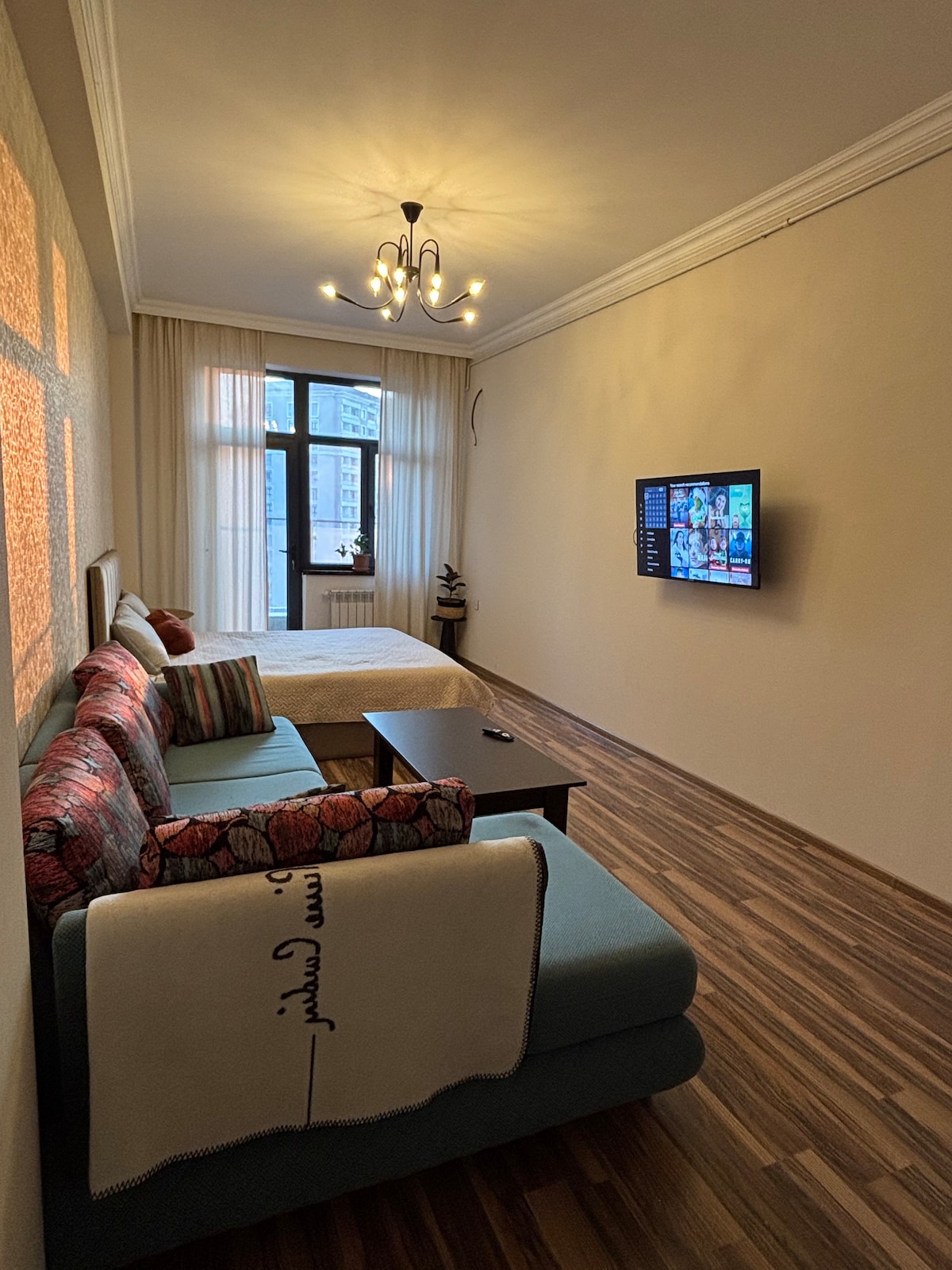 A bright studio space showcases a spacious bed and a comfortable sofa bed, accompanied by a dark coffee table. A wall-mounted television is visible, and sheer curtains allow natural light to enter. The wooden flooring enhances the overall appearance of the room.