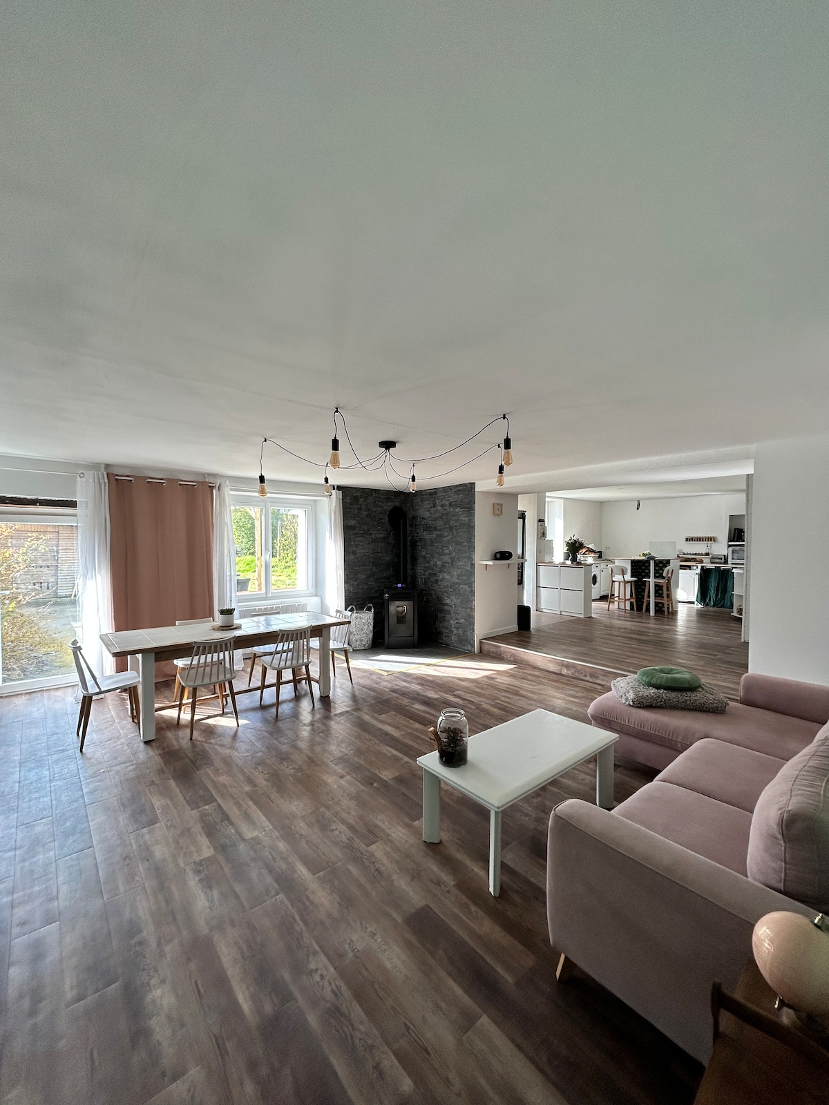 An open living space is showcased, featuring a cozy seating area with a soft sofa and a simple coffee table. A dining table for six is positioned next to large windows, allowing natural light to illuminate the room. A wood stove contributes to the ambiance.