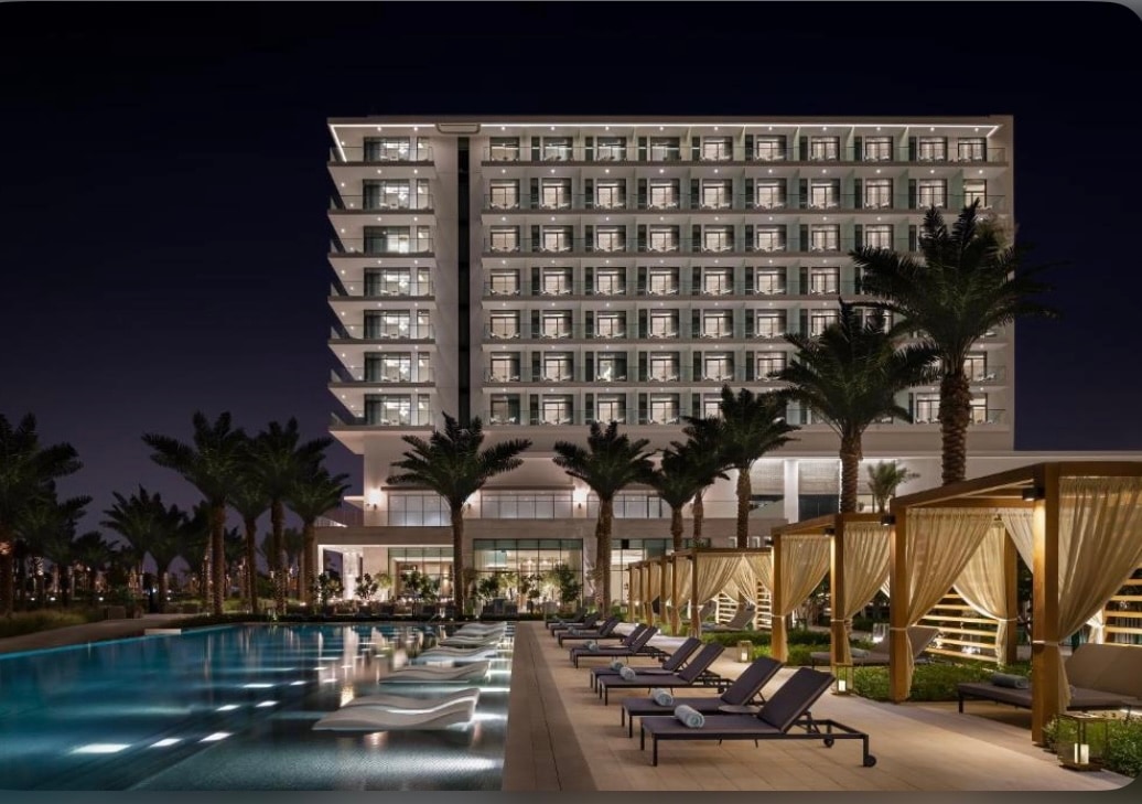 A modern hotel structure is presented at night, featuring multiple levels of illuminated windows. A swimming pool is visible in the foreground, lined with lounge chairs and shaded cabanas, complemented by palm trees that add to the tranquil atmosphere.