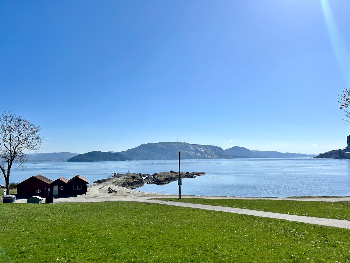 A serene waterfront scene features clear blue skies and calm waters, with gentle hills visible in the distance. Red storage buildings are positioned near the shoreline, and a grassy area invites leisurely strolls along the water's edge.