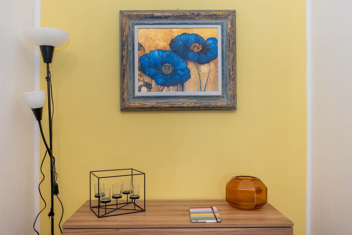 A vibrant yellow wall highlights a floral artwork featuring blue flowers in an ornate frame. Below, a wooden side table presents a decorative glass vase and a geometric candle holder, complemented by a colorful notepad and pen arrangement.