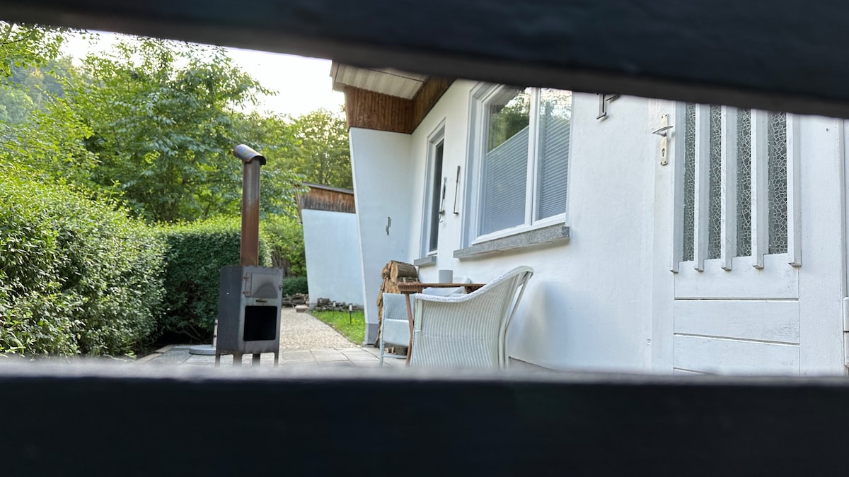 A view is offered through black slats, revealing a charming outdoor area. A white chair and small table are placed beside a solid stove, surrounded by greenery. The exterior of the house is visible, featuring large windows and a welcoming entrance.