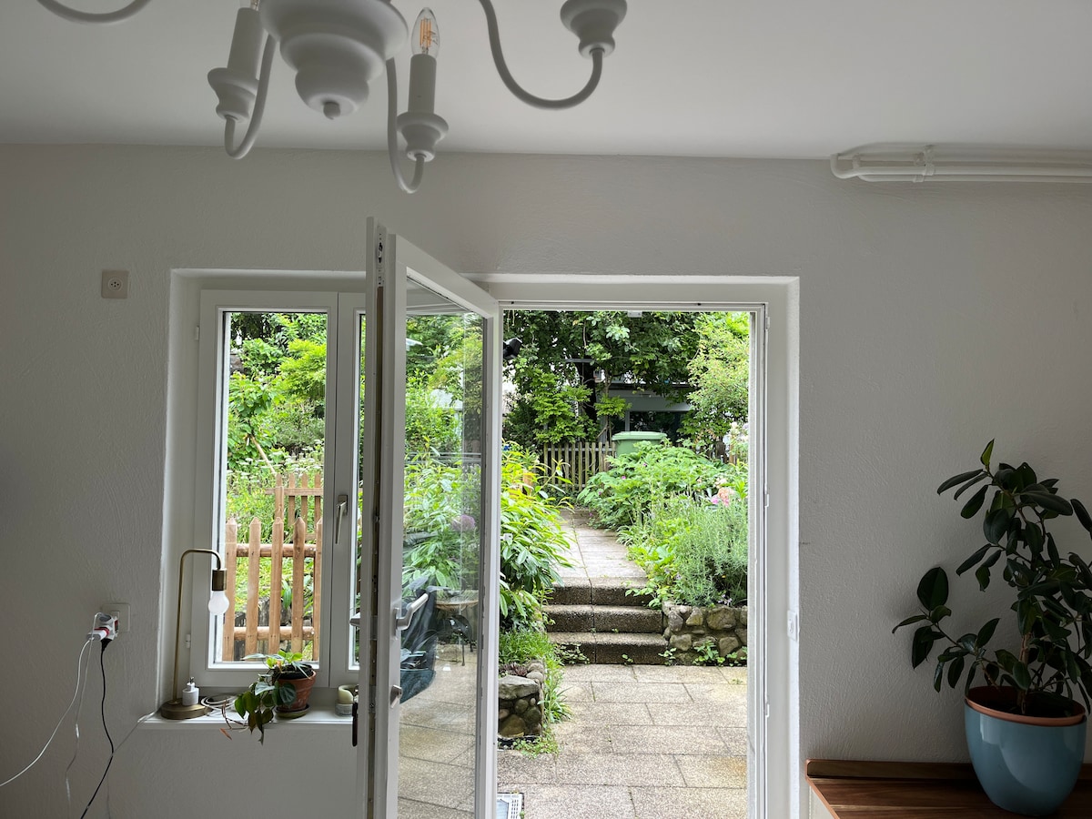 The image displays an open door leading to a lush garden, framed by two windows. The pathway is lined with greenery, inviting a sense of tranquility. A light fixture hangs from the ceiling, while potted plants are situated nearby, adding a touch of nature indoors.