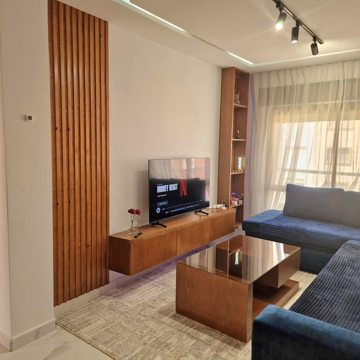 A modern living area features a dark blue sectional sofa and a sleek wooden media console. A flat-screen TV is mounted on the wall, and a glass coffee table sits on a light rug. Natural light filters through sheer curtains covering large windows.