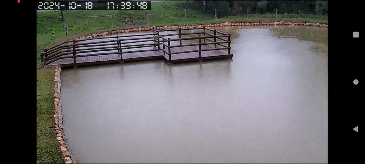 A wooden deck is positioned over a calm body of water, creating a serene setting. Surrounding greenery is visible in the background, offering a natural atmosphere. The water appears slightly murky, indicating recent rainfall, while the deck's railings provide a rustic touch.