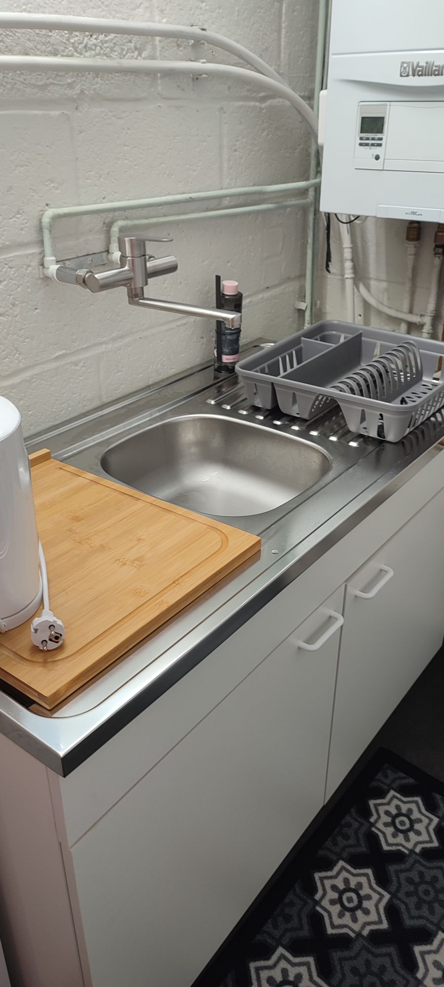 A compact kitchen area features a metallic sink with a modern faucet. A wooden cutting board rests on the countertop, next to a drying rack with dishes. Below, cabinetry provides storage, while plumbing fixtures are visible on the wall.