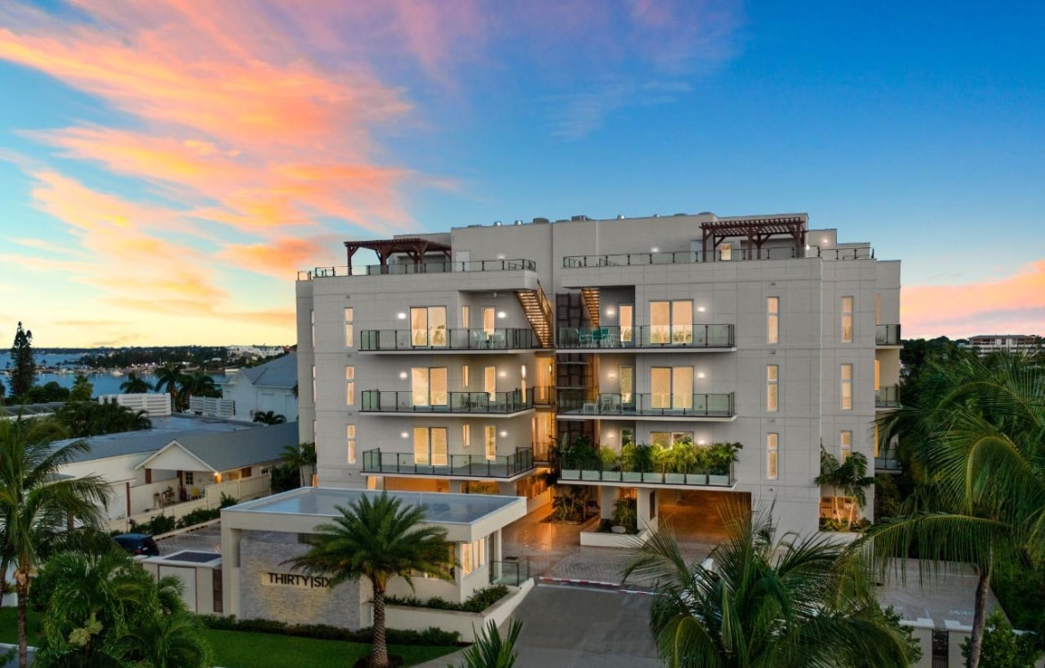 A modern penthouse building stands amidst lush palm trees, featuring multiple balconies with large glass doors and inviting outdoor spaces. The sunset sky casts a warm glow, accentuating the clean lines and contemporary design of the structure.