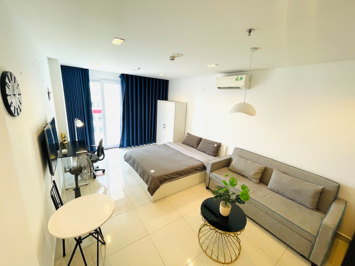 A modern studio features a spacious bed with neutral bedding and a cozy sofa. An office chair sits at a small table, while a desk with a monitor is also present. Large windows are dressed with dark curtains, and a small plant adds a touch of greenery.