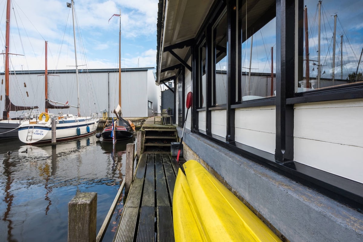 A view of the wooden dock alongside the water, featuring two yellow kayaks ready for use. Reflections of the surrounding boats and buildings are visible on the calm water surface, creating a serene atmosphere.