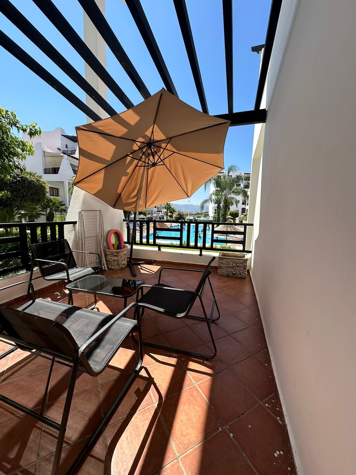 A spacious terrace is designed with black railings and offers a seating area with four chairs arranged around a small table. A large beige umbrella provides shade, while sunlight casts patterns on the tiled floor. A view of the pool area is visible in the background.