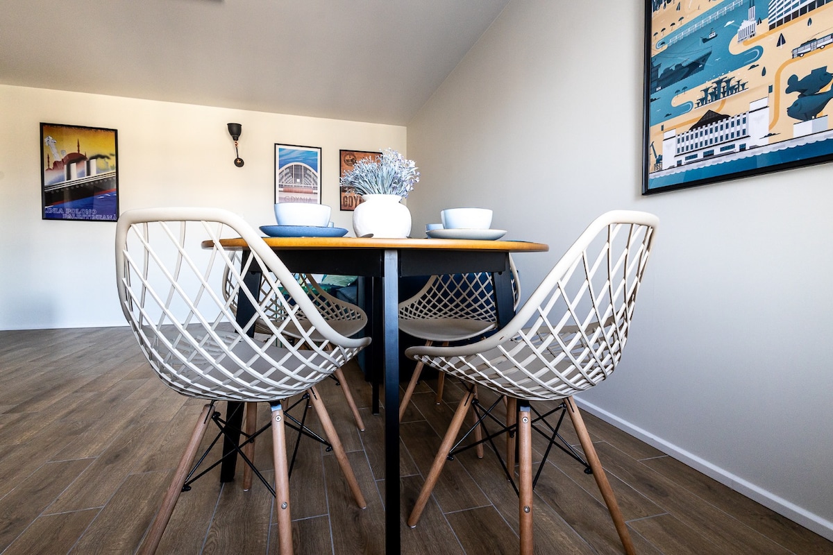 A dining area features a wooden table surrounded by four white wire chairs with wooden legs. On the table, a decorative arrangement of flowers is paired with plates and bowls. Walls are adorned with framed art depicting coastal scenes.