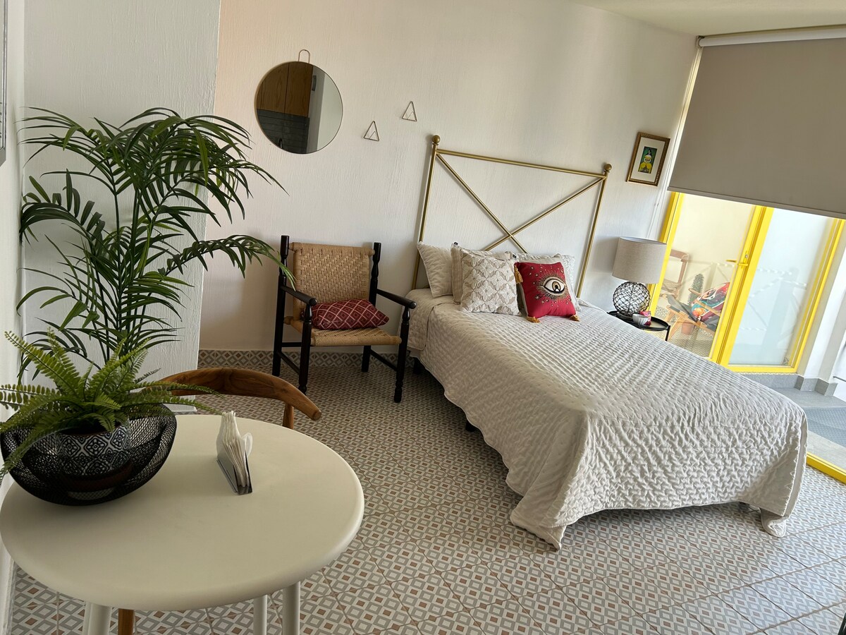 A bright and airy sleeping area is furnished with a spacious bed, accented by a colorful decorative pillow. A small table with a plant and a wooden chair are positioned nearby, while large windows allow ample natural light into the room.