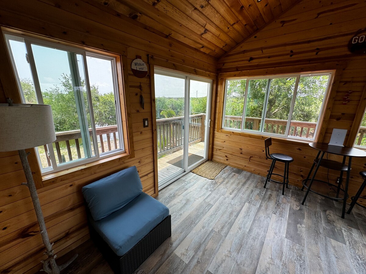 A bright seating area features large windows that invite natural light, showcasing views of the surrounding landscape. A comfortable blue chair rests beside a small table with two black stools, while a lamp adds a cozy touch. Wooden walls and flooring enhance the cabin's ambiance.