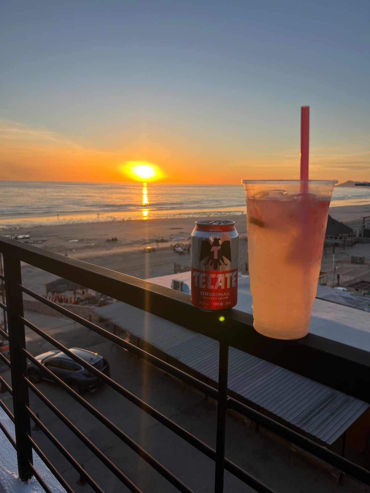 A sunset view is captured from a private balcony, featuring a refreshing drink in a clear cup alongside a canned beverage. The beach and ocean are visible below, with waves gently lapping at the shore as the sun descends behind the horizon.