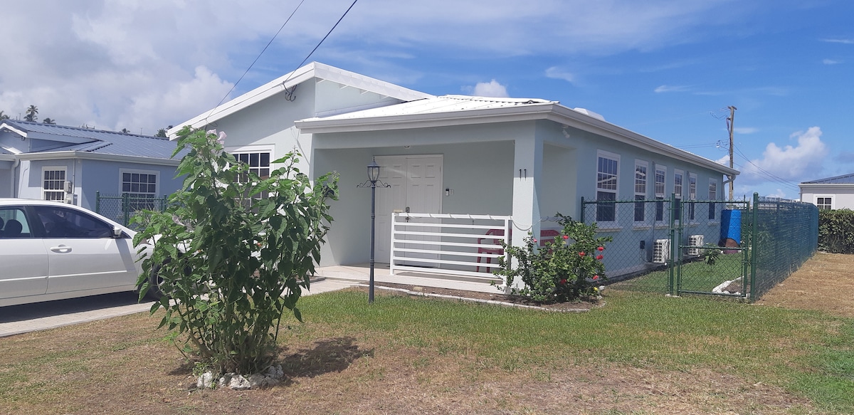 A light blue house features a covered entrance with a white door, framed by decorative fencing and a small garden area. A car is parked in the driveway, and a well-maintained lawn is visible in the foreground, providing a welcoming view of the property.