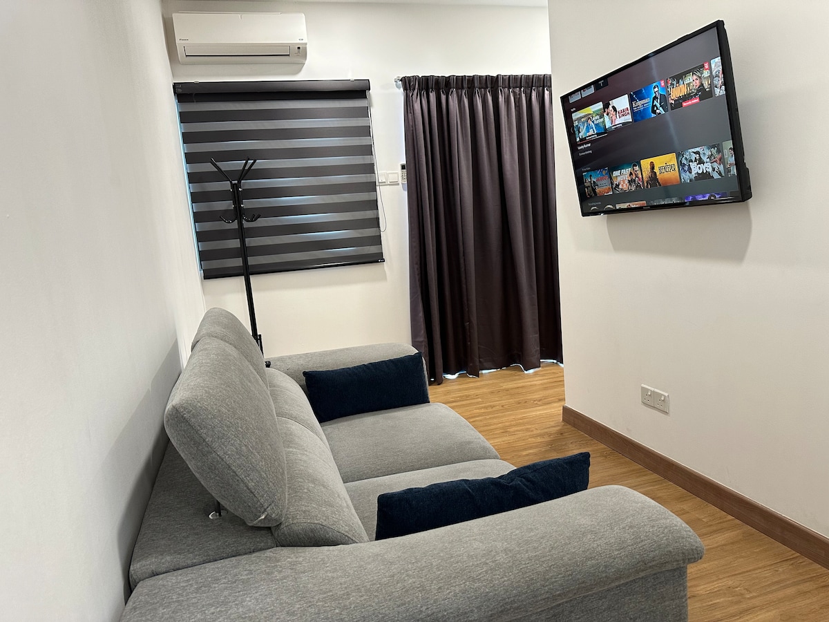A spacious corner sofa in muted grey is positioned near a wall-mounted TV. The room is illuminated by natural light filtered through fitted dark blinds. A set of dark curtains adds privacy, while a coat rack stands against the wall.