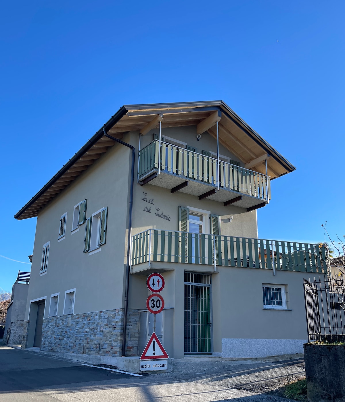 The three-story building features a light facade with wooden accents and a welcoming balcony. Green shutters frame the windows, and a stone base adds texture. Street signs are visible in the foreground, indicating traffic regulations.
