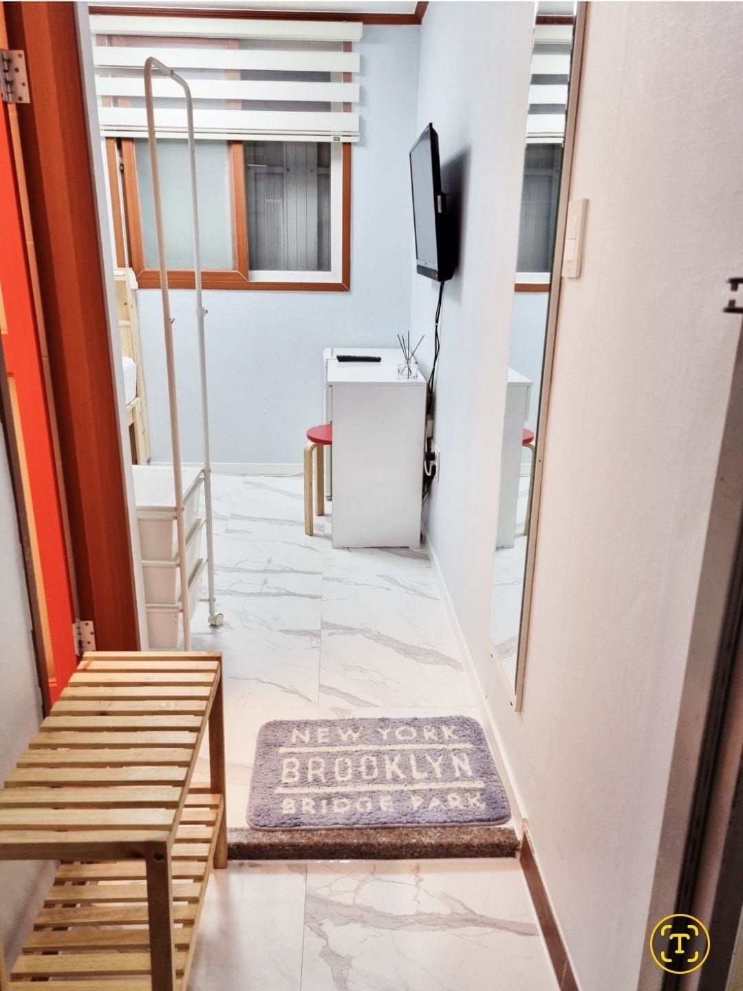 A hallway area is shown, featuring a small white desk with two red chairs positioned nearby. A wall-mounted television is visible across from a standing mirror. A welcome mat with an urban theme lies on the tiled floor, and natural light is filtered through window treatments.