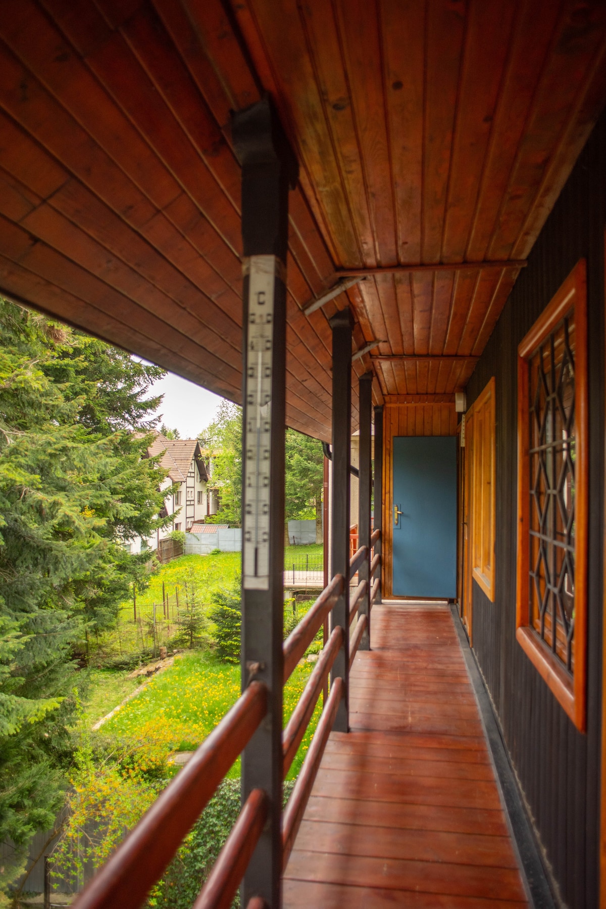 A covered wooden balcony extends along the exterior, framed by dark walls and wooden beams. A solid blue door is visible at the end of the corridor, while greenery and a view of the neighboring property can be seen through the railing.