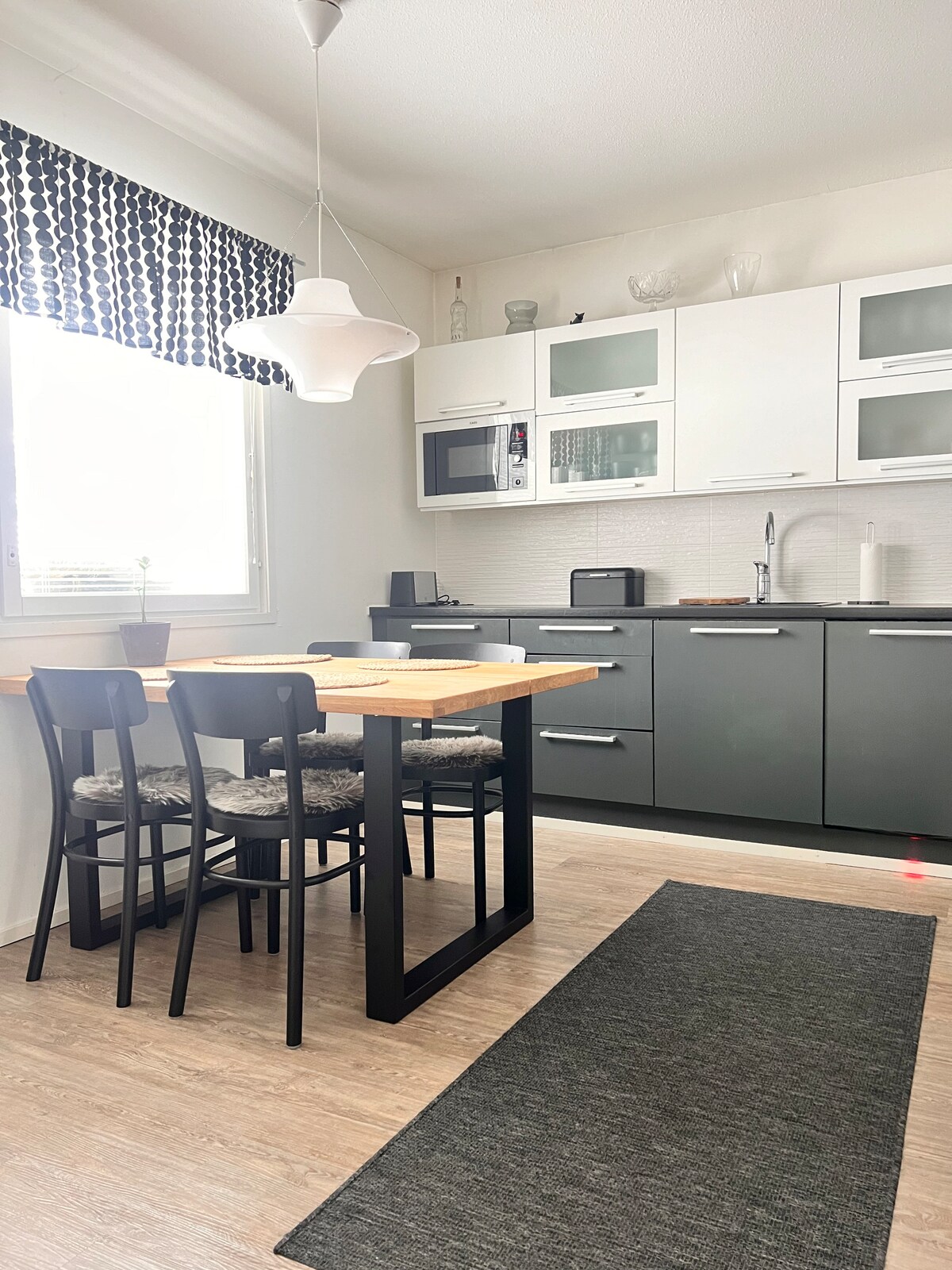 A modern kitchen area is displayed, featuring sleek gray cabinetry and a light wood dining table. Three black chairs are arranged around the table. Natural light streams in through a window adorned with patterned curtains, while essential appliances are neatly integrated within the cabinetry.