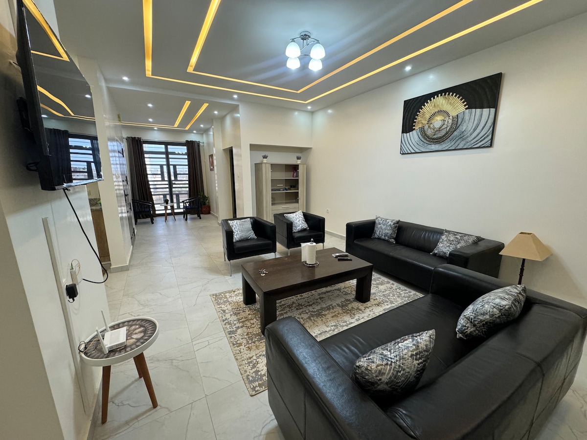 A modern living area features black leather sofas arranged around a central coffee table. Soft lighting highlights the sleek design, while a large artwork adorns the wall. Natural light enters through expansive windows, enhancing the spacious and inviting environment.
