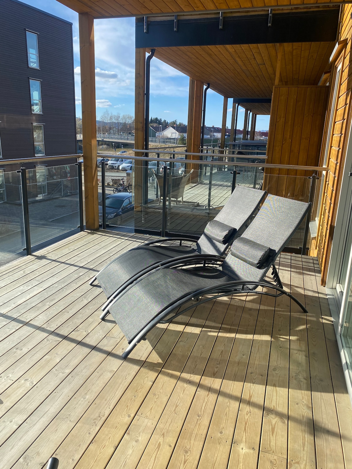 A wooden balcony features two comfortable lounge chairs arranged for relaxation. The floor is made of smooth wood planks, and a glass railing provides an unobstructed view. Bright sunlight fills the space, enhancing the open feel, with nearby buildings visible in the background.
