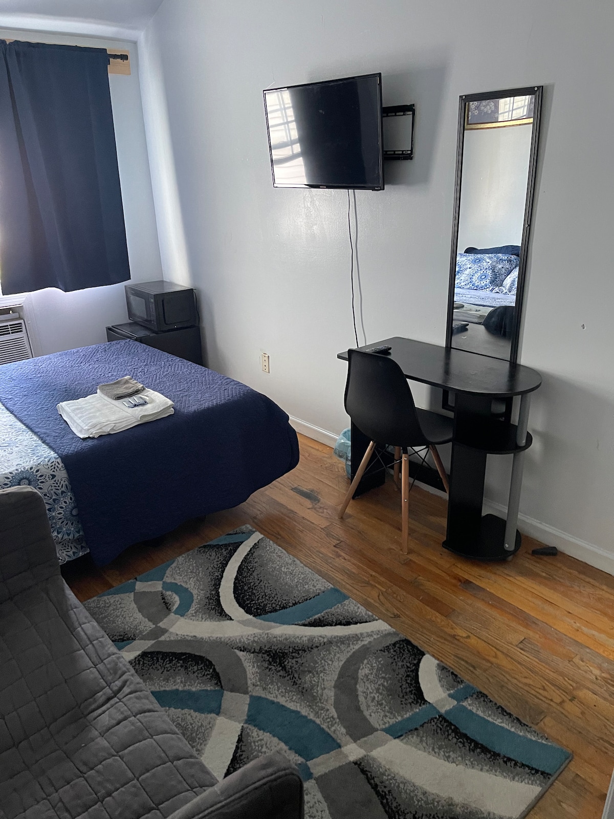The room features a cozy sleeping area with a blue and white patterned blanket covering the bed. A flat-screen TV is mounted on the wall opposite. A mirror and a desk chair are positioned nearby. Natural light enters through a window, partially obscured by dark curtains.