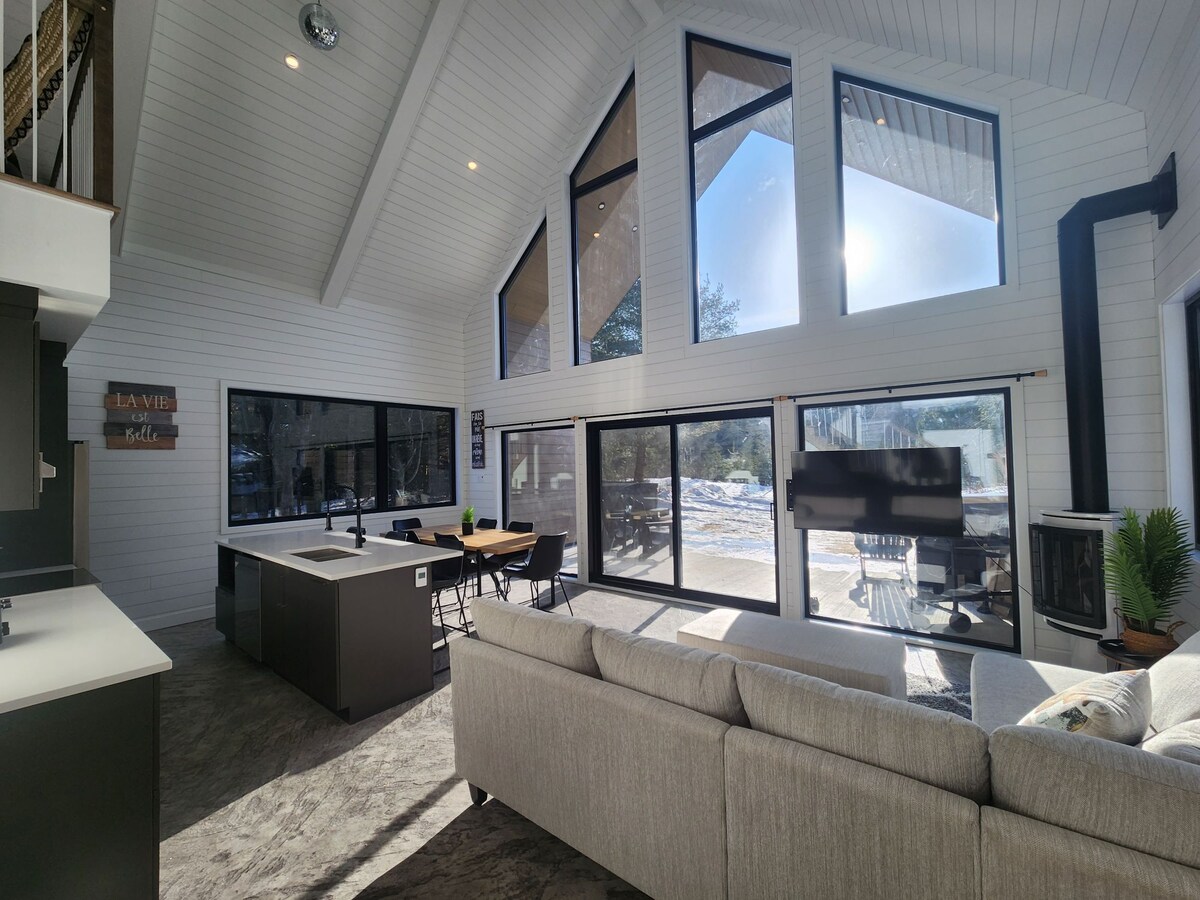 A spacious living area is highlighted by large floor-to-ceiling windows, allowing natural light to fill the space. A modern sectional sofa faces a sleek entertainment unit. An open kitchen area features a large island, complemented by a dining table with chairs.