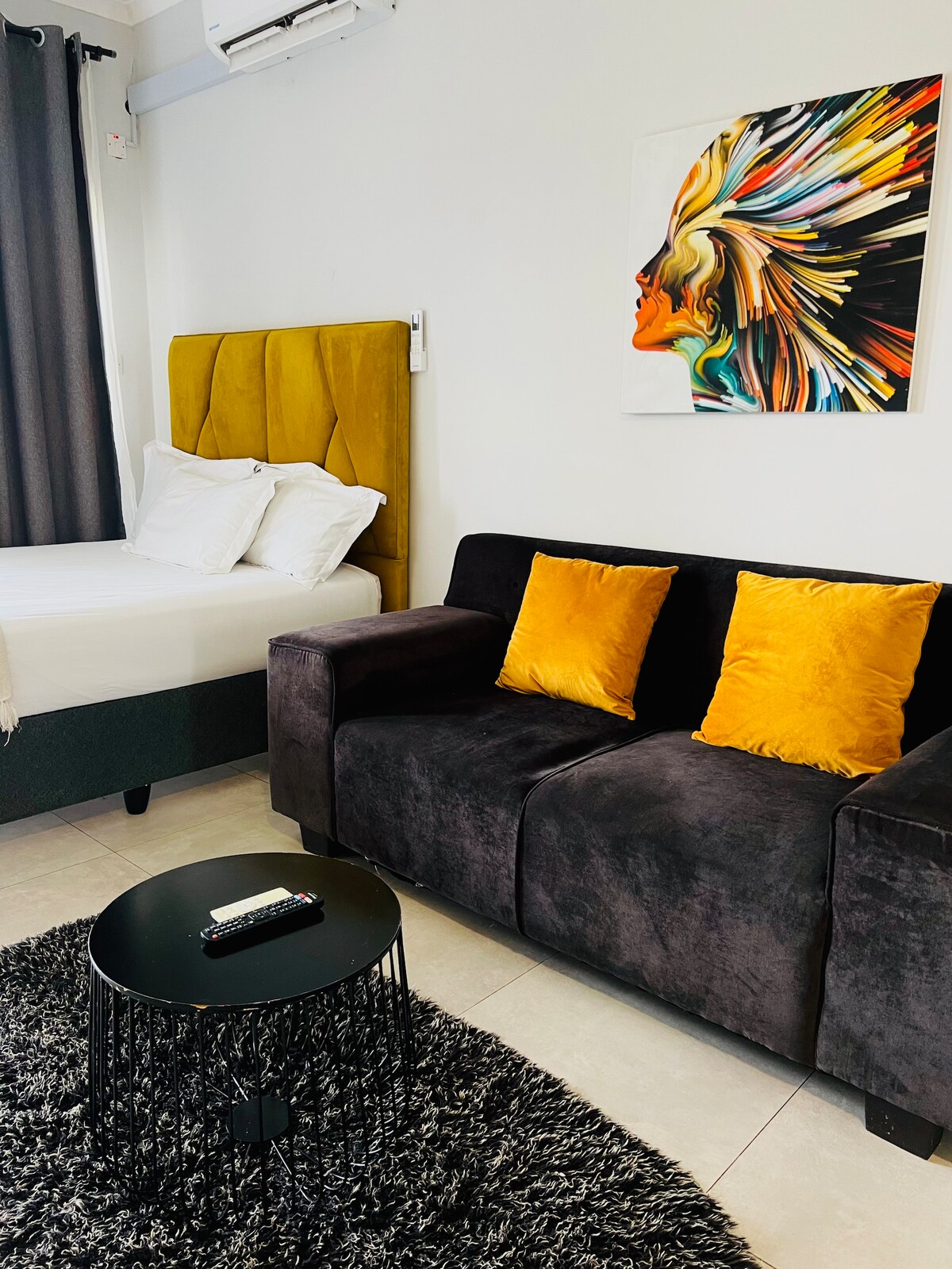 A cozy studio apartment features a spacious bed with a yellow upholstered headboard, complemented by crisp white linens. A dark couch adorned with two mustard-colored pillows sits opposite a circular black coffee table on a textured rug. A colorful wall art piece adds visual interest.