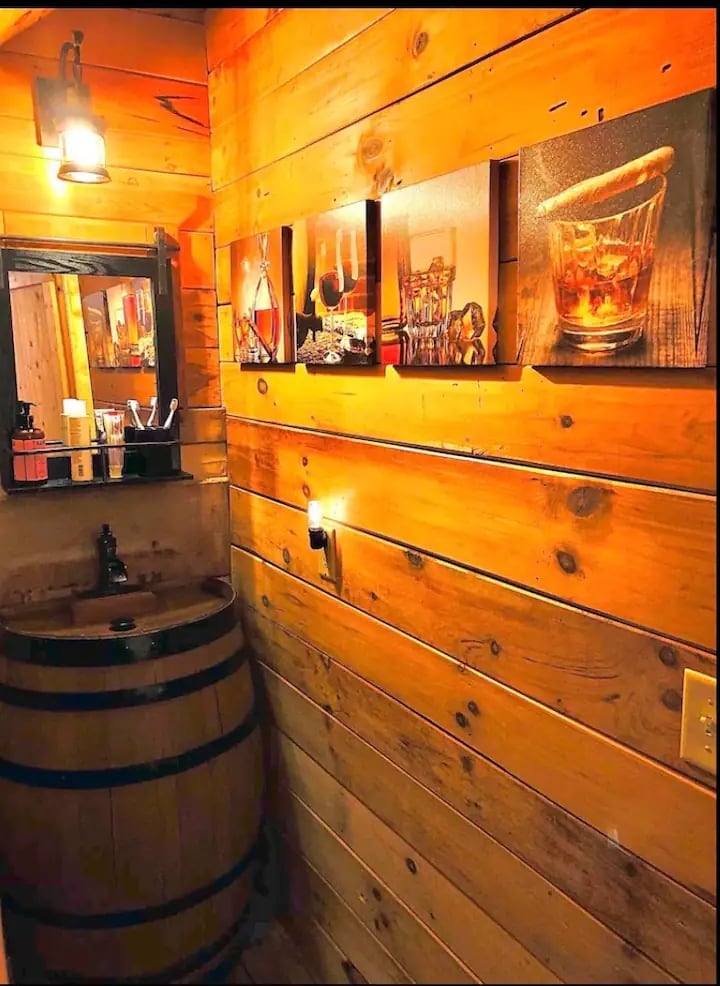 A warm and inviting space features wooden walls and a decorative arrangement of three framed images related to drinks. A rustic barrel serves as a base for a sink, enhancing the cabin's charm. Soft lighting illuminates the area, creating a cozy ambiance.