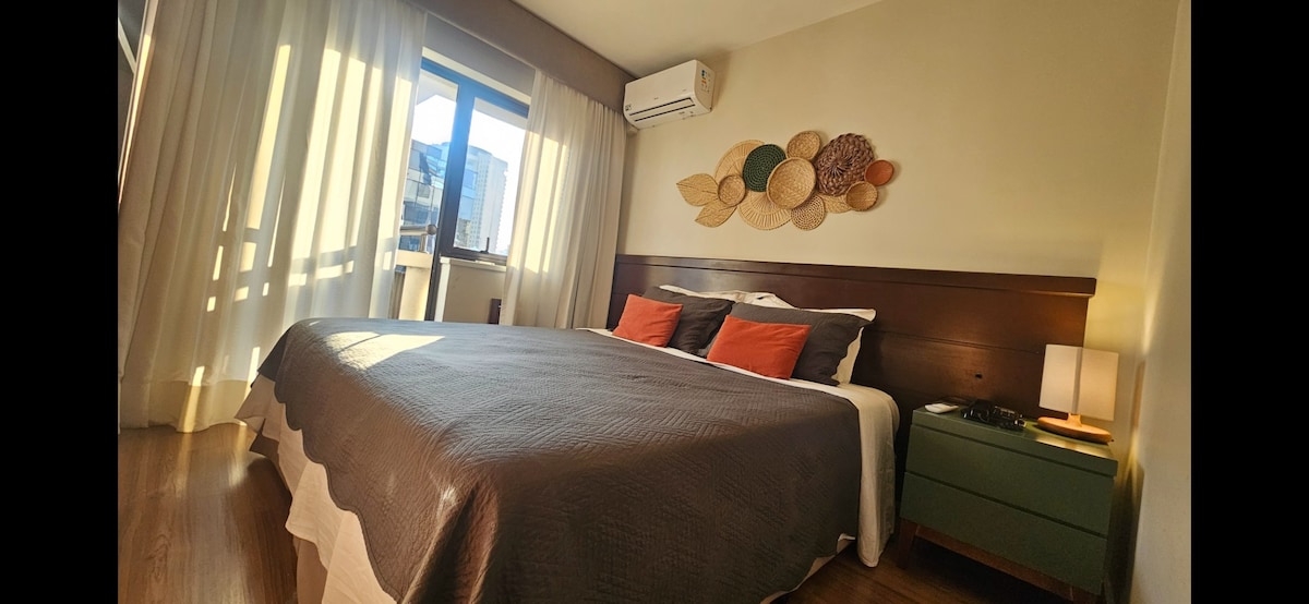 A comfortable bedroom features a spacious bed with decorative pillows, complemented by warm wooden accents. Natural light pours through a large window, illuminating the soft curtains. Wall decor adds a touch of texture, enhancing the overall ambiance of the space.