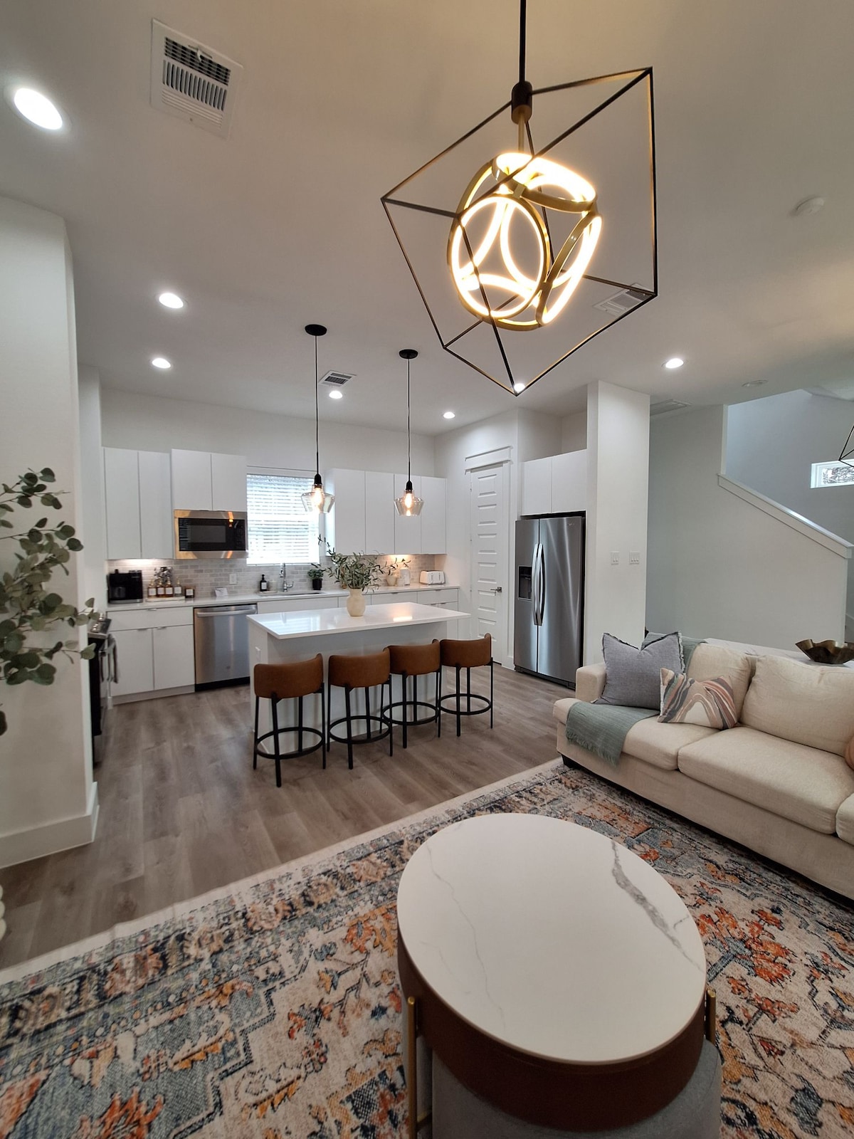 An open-concept living area showcases a modern kitchen with sleek cabinetry and stainless steel appliances. A large island with four bar stools provides additional seating. Natural light fills the space, highlighting a cozy lounge area with a light-colored sofa and decorative accents.