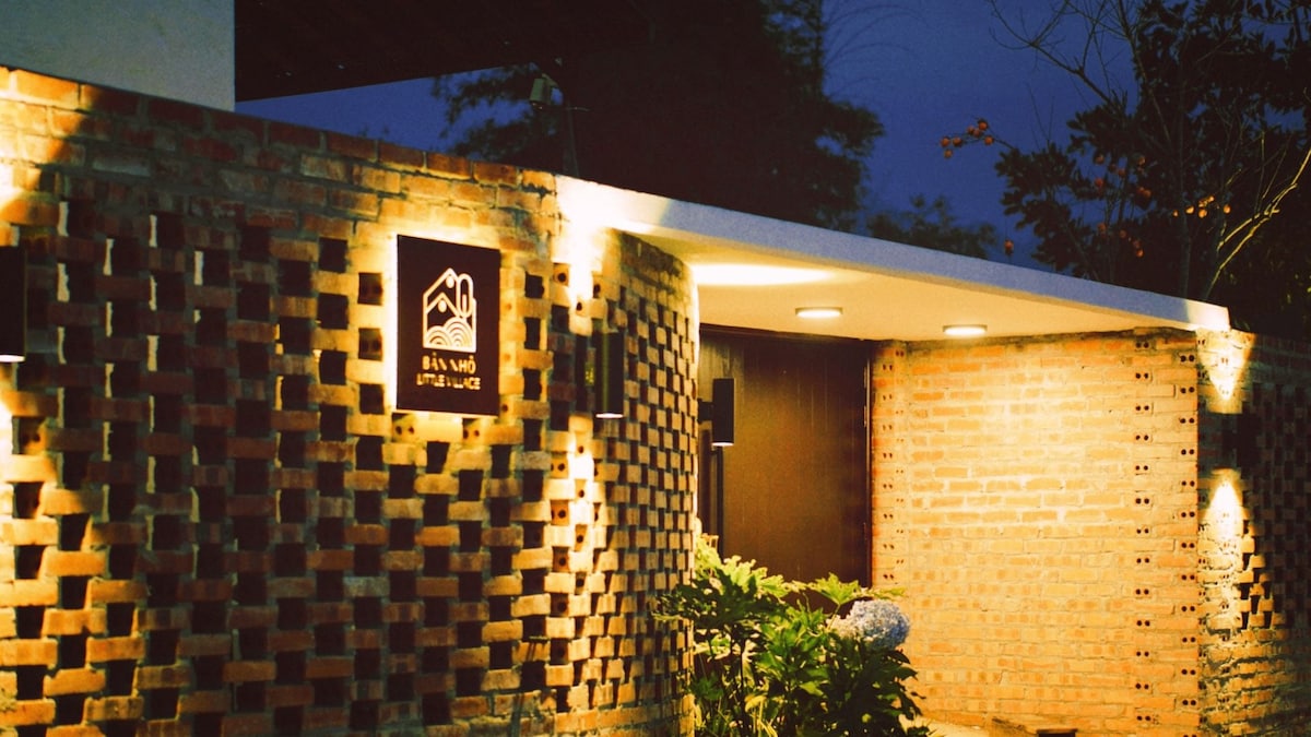 The entrance of the building is framed by textured brick walls, illuminated by strategically placed lighting. A welcoming sign is displayed prominently, while lush greenery adds natural beauty to the surroundings.
