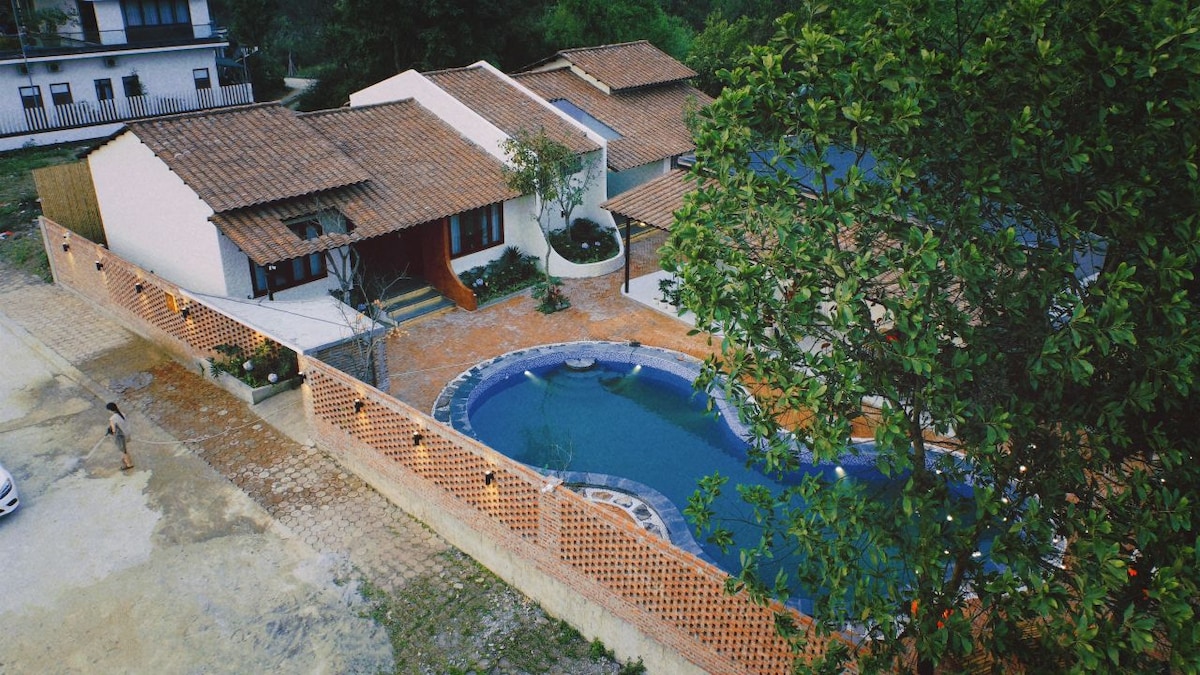 The image captures a bird's-eye view of a property featuring a curved outdoor pool surrounded by a spacious tiled area. The traditional-style buildings with terracotta roofs are visible, and lush greenery partially frames the scene, enhancing the inviting outdoor experience.