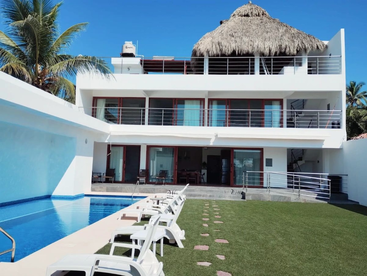 The modern villa features a thatched roof and a spacious outdoor area with a landscaped lawn. A private pool is situated adjacent to the building, with lounge chairs positioned along the water's edge. Large glass doors provide access to the interior living spaces.