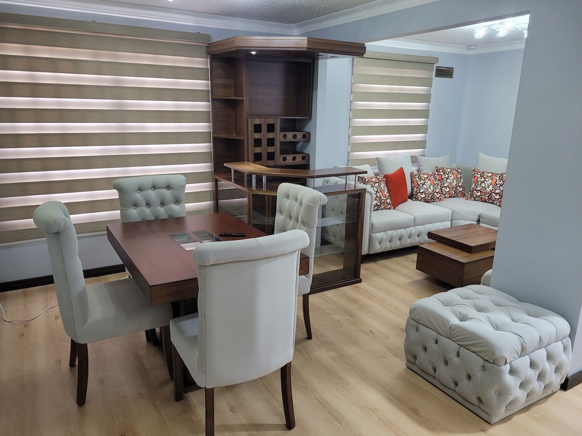 A modern living space features a wooden dining table accompanied by upholstered chairs and a comfortable seating area with decorative cushions. A glass display cabinet and a coffee table complete the arrangement, while soft lighting enhances the contemporary design.