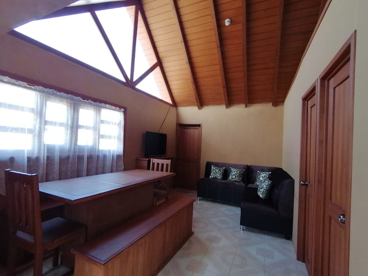 A cozy living area is presented, featuring a textured wooden ceiling with triangular beams. A wooden dining table is paired with four chairs, while a dark sofa adorned with decorative pillows is positioned against the wall. Windows allow natural light to filter through the sheer curtains.