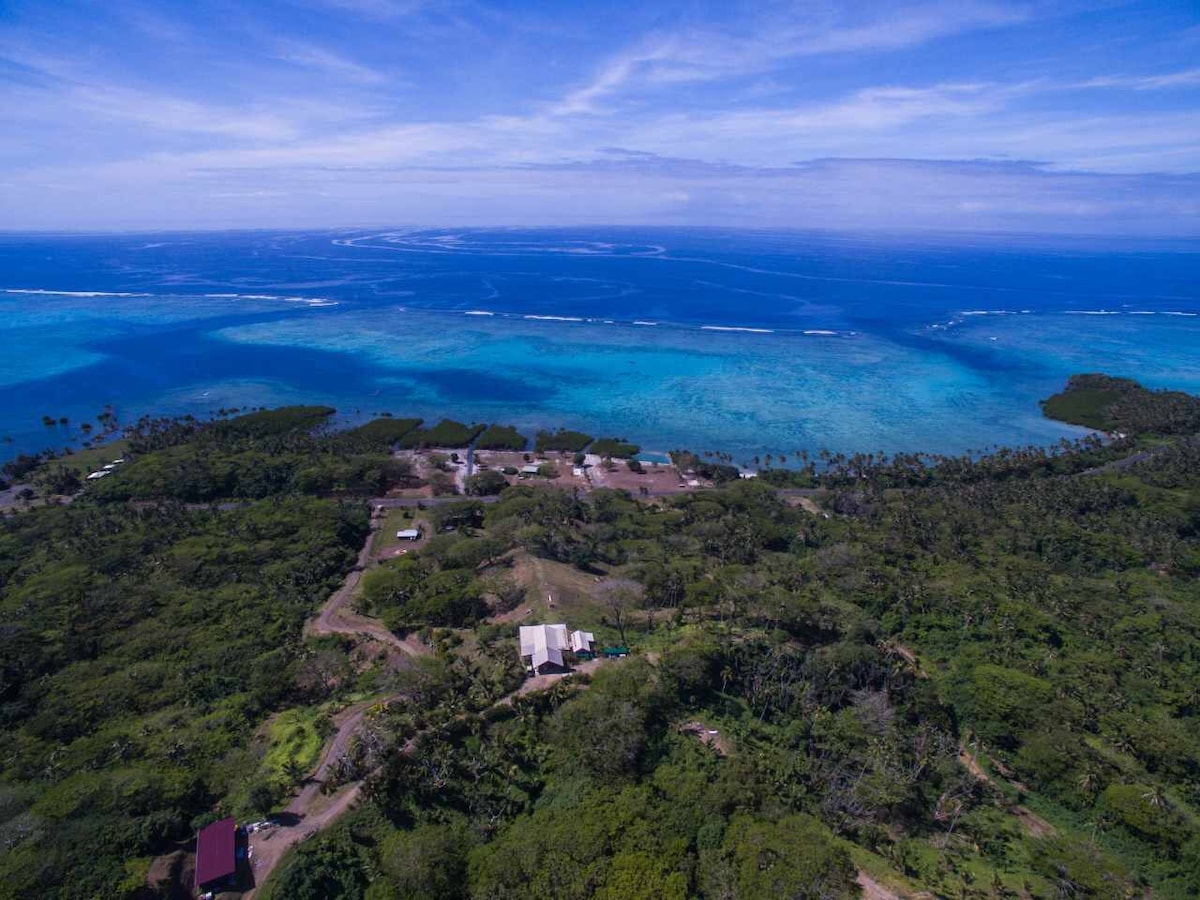 An aerial view showcases the vibrant blue waters of the ocean, contrasting with lush green land. The layout includes various terrains leading to the coastline, with scattered structures blending into the natural landscape, offering a sense of seclusion and tranquility.