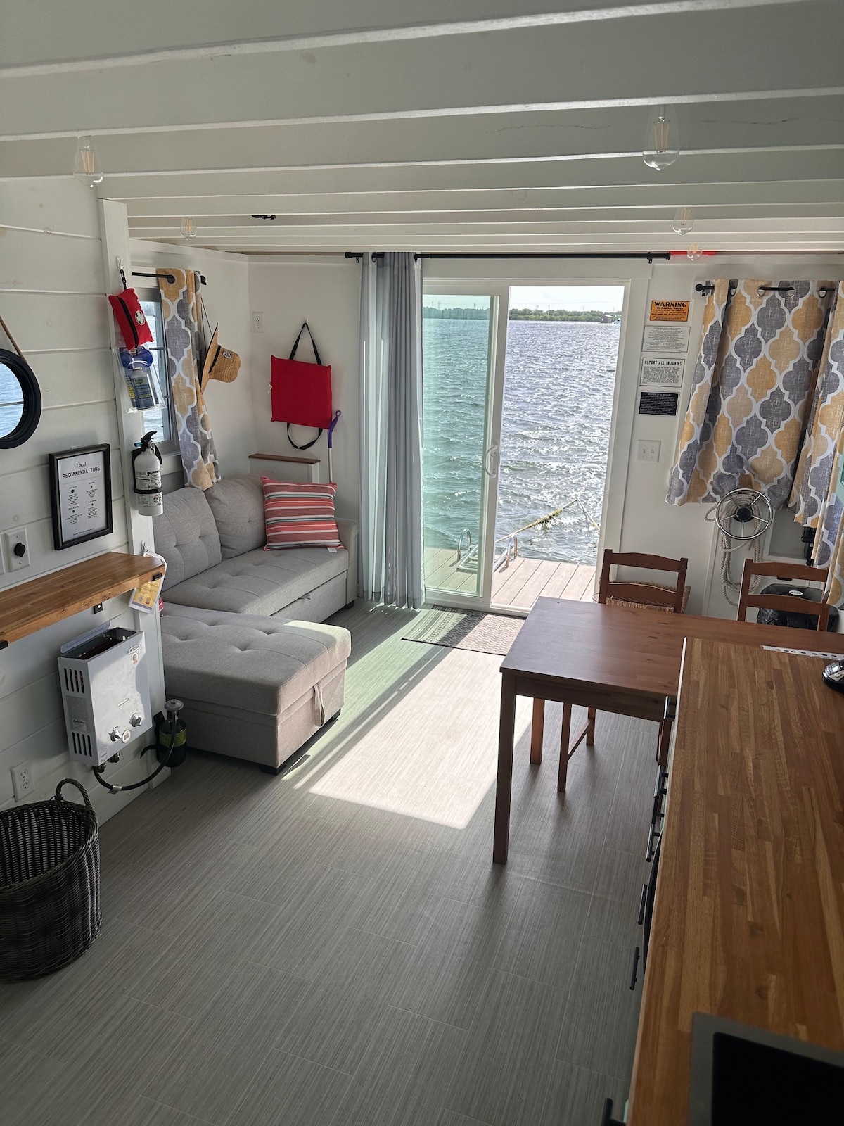 The living area features a comfortable sofa positioned beside large windows, allowing natural light to fill the space. A small table and chairs are present, with decorative curtains framing the view of the water just outside. The entrance leads to a wooden deck overlooking the serene bay.
