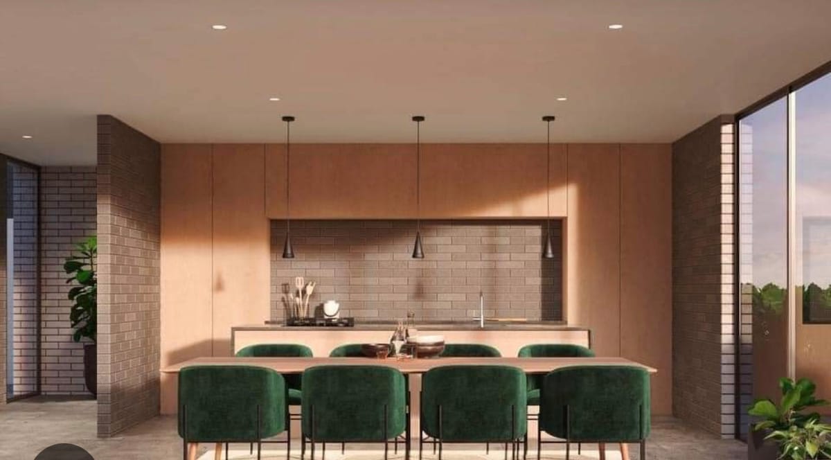 A contemporary kitchen area showcases a long wooden dining table surrounded by plush green chairs. Minimalist pendant lights hang from the ceiling, illuminating the space, while a sleek kitchen counter is visible in the background, featuring modern appliances and a tiled backsplash.