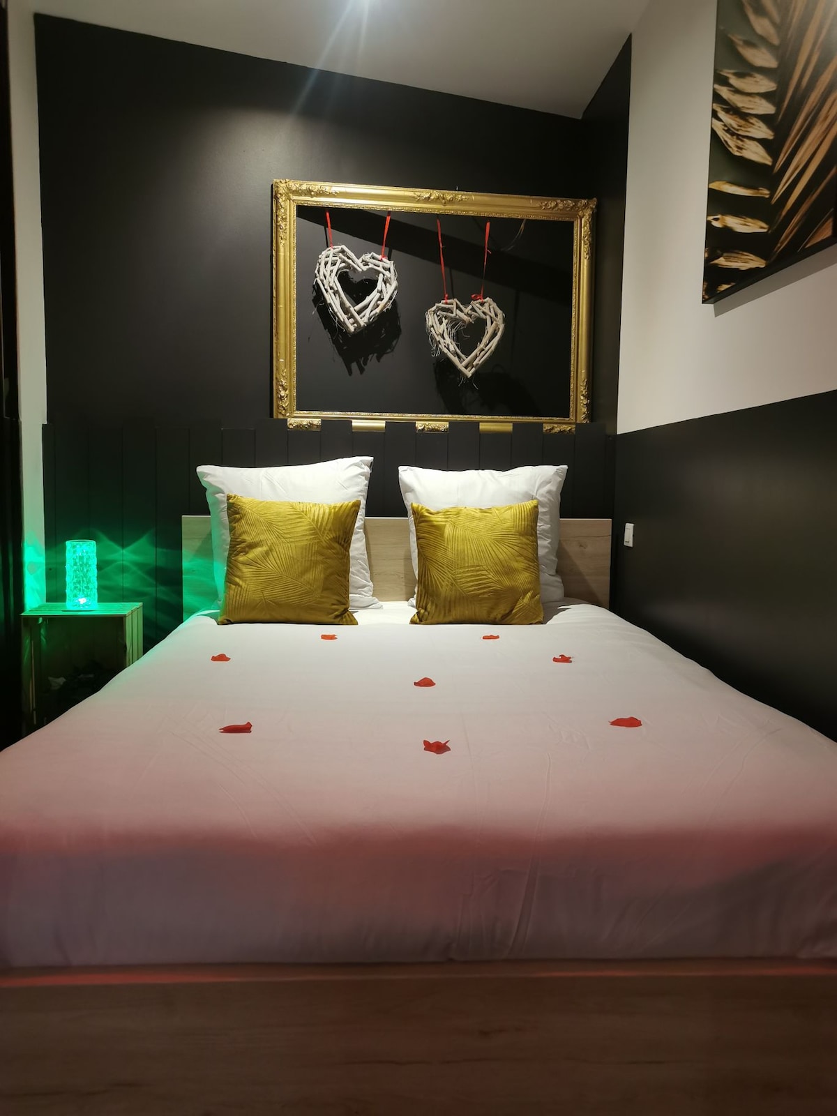 A neutral-toned sleeping area features a spacious bed adorned with rose petals. Two decorative pillows in gold are positioned at the head of the bed. Framed heart decorations are displayed above the bed, complemented by ambient green lighting from a nearby lamp.