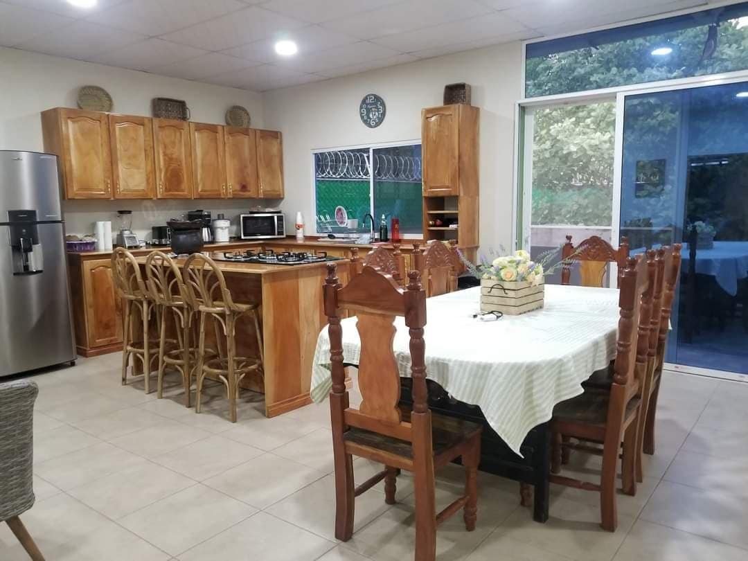 A spacious kitchen and dining area features wooden cabinetry and a large table surrounded by chairs. Modern appliances, including a refrigerator and microwave, are visible. Natural light enters through the sliding glass doors, illuminating the room's earthy tones.