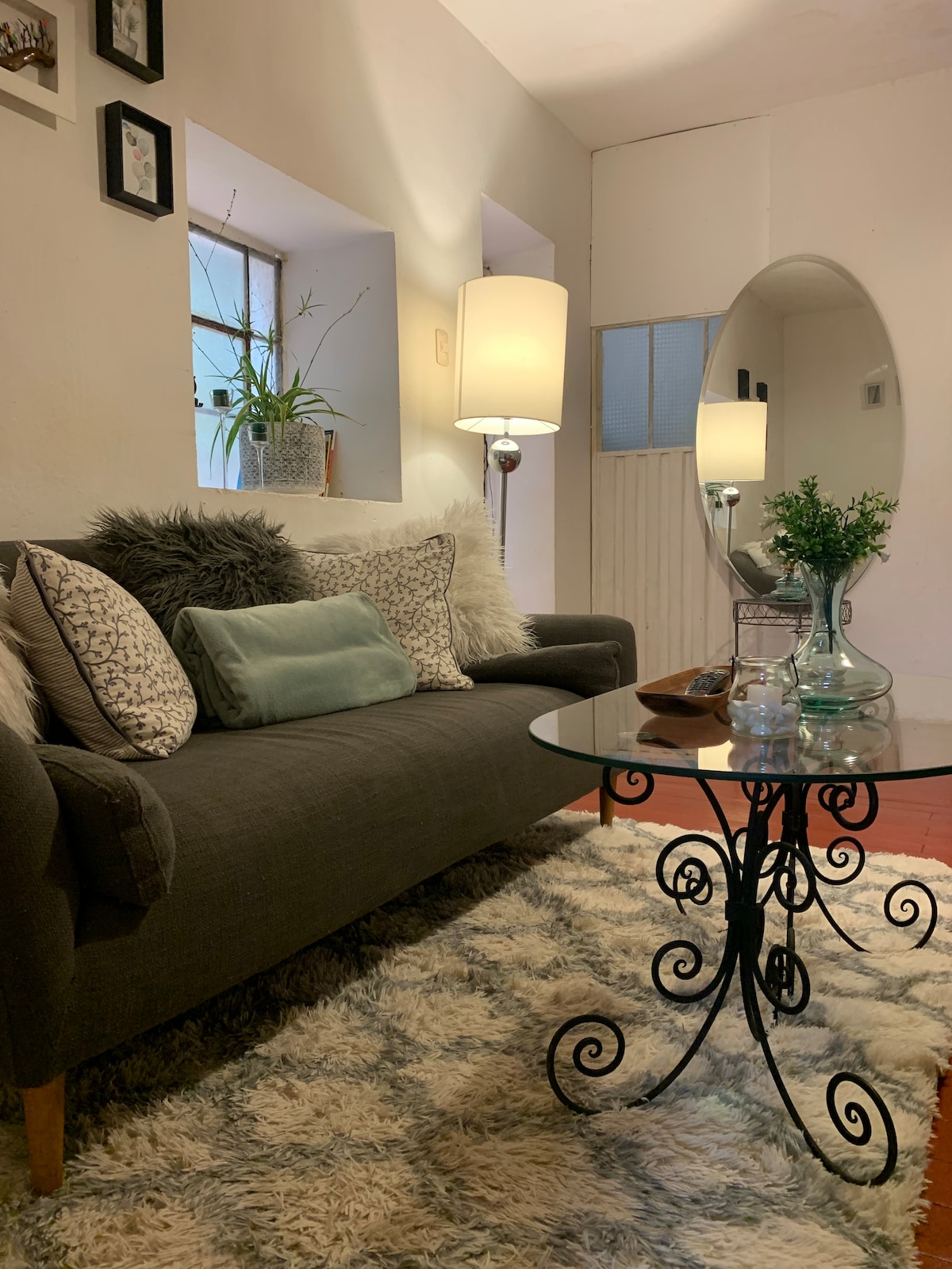 A cozy living area features a plush gray sofa adorned with decorative pillows. A glass table with an ornate black base sits on a soft, textured rug. Natural light enters through a nearby window, illuminating the room alongside a standing lamp.