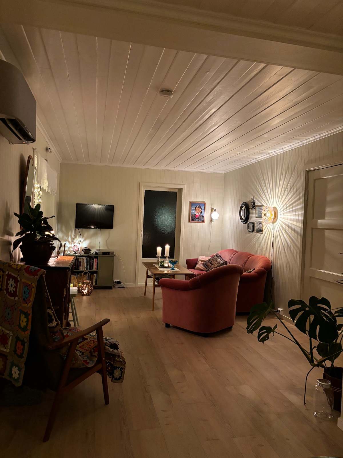 A cozy living area features a pink armchair and a small side table, softly illuminated by candles. A television is mounted on the wall, and decorative pieces are displayed on a nearby cabinet. Natural light reflects off a glass door leading to another room.