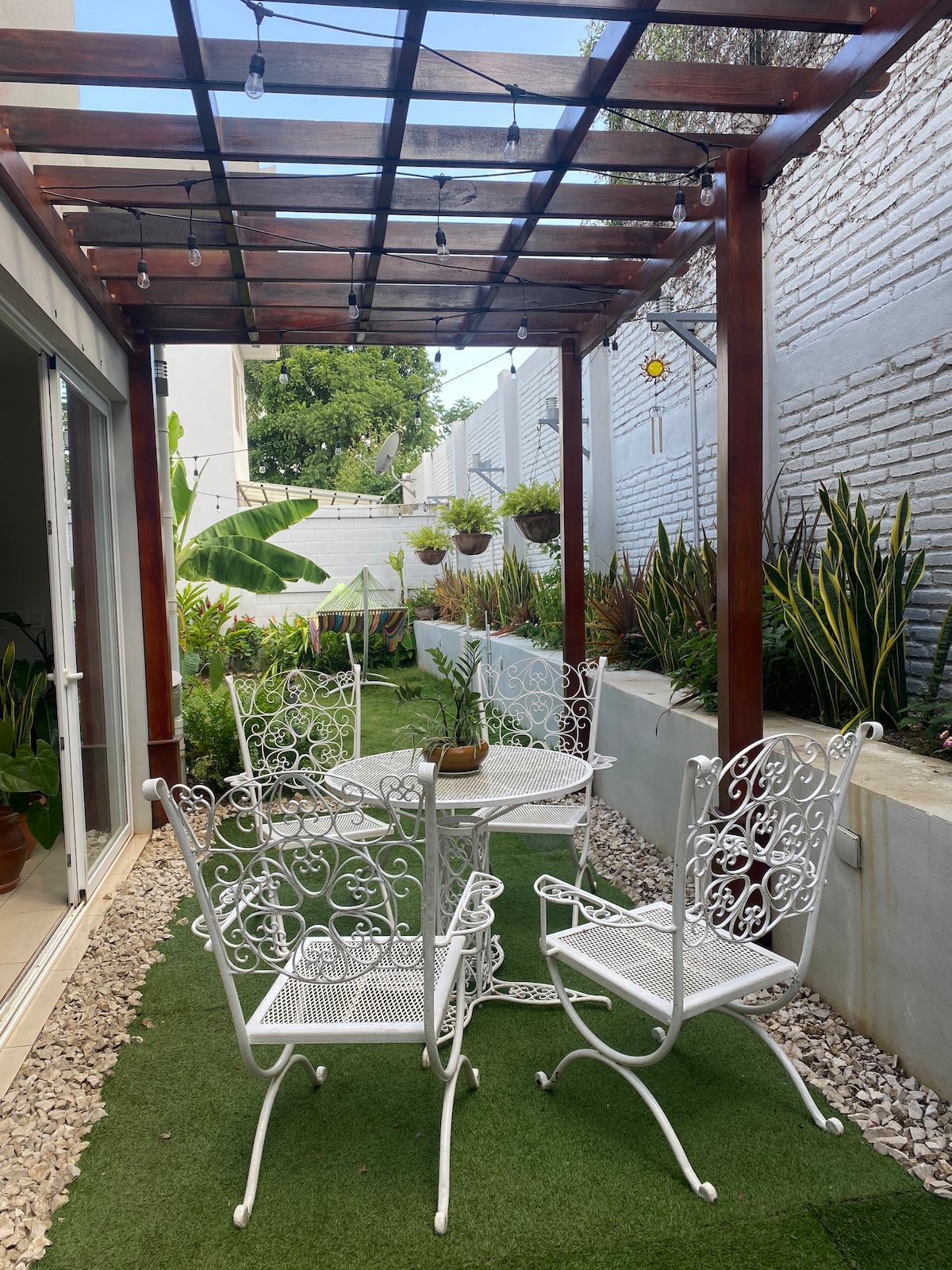 An outdoor seating area is presented with a white metal table and four ornate chairs under a wooden pergola. The space features lush greenery and hanging plants, along with decorative lighting overhead, creating a serene environment for relaxation.