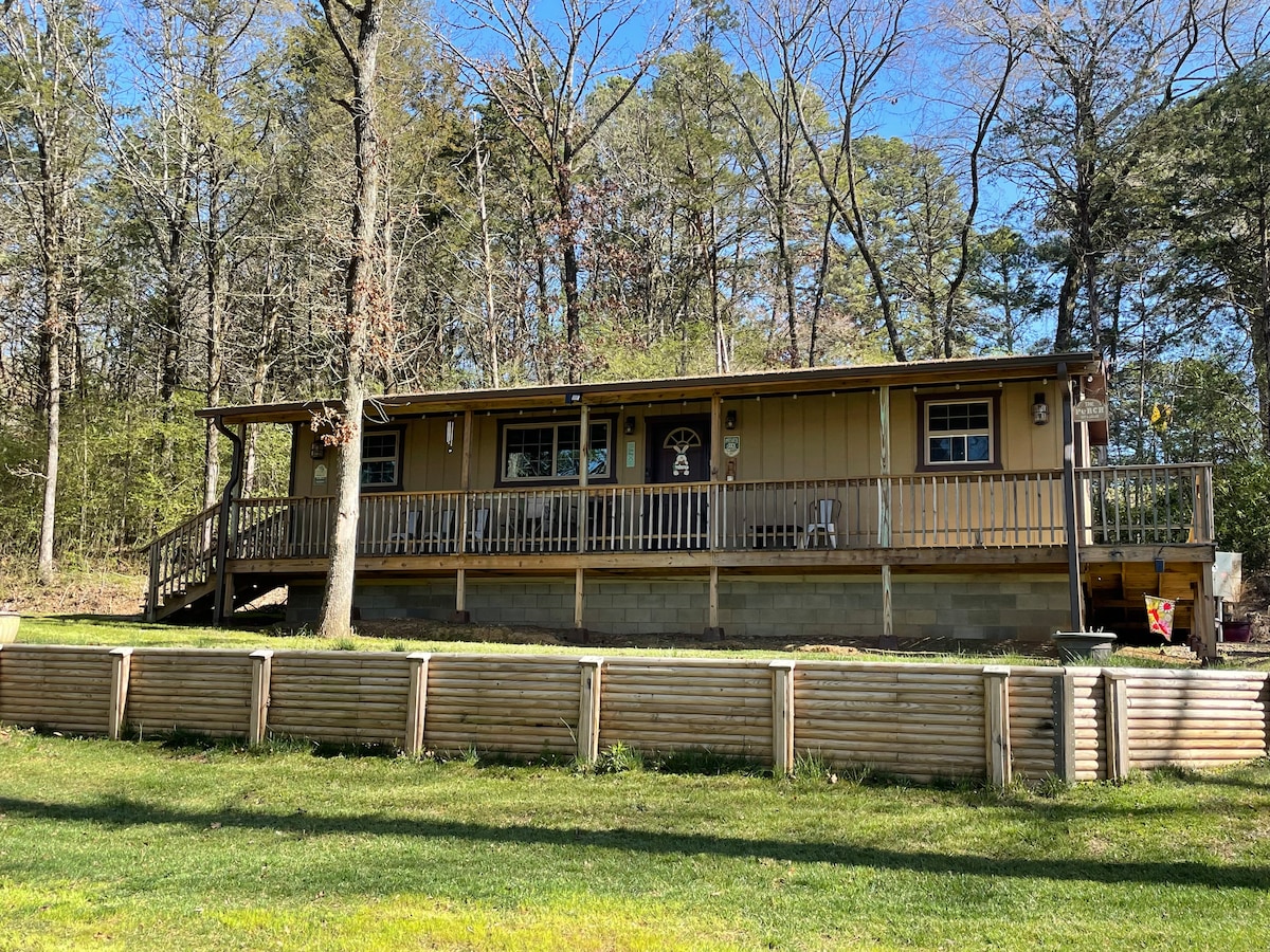 A wooden cabin is set amidst trees, displaying a welcoming front porch with a railing. Sunlight filters through the surrounding greenery onto a grassy area in the foreground, while the structure showcases multiple windows and a door with a decorative feature.