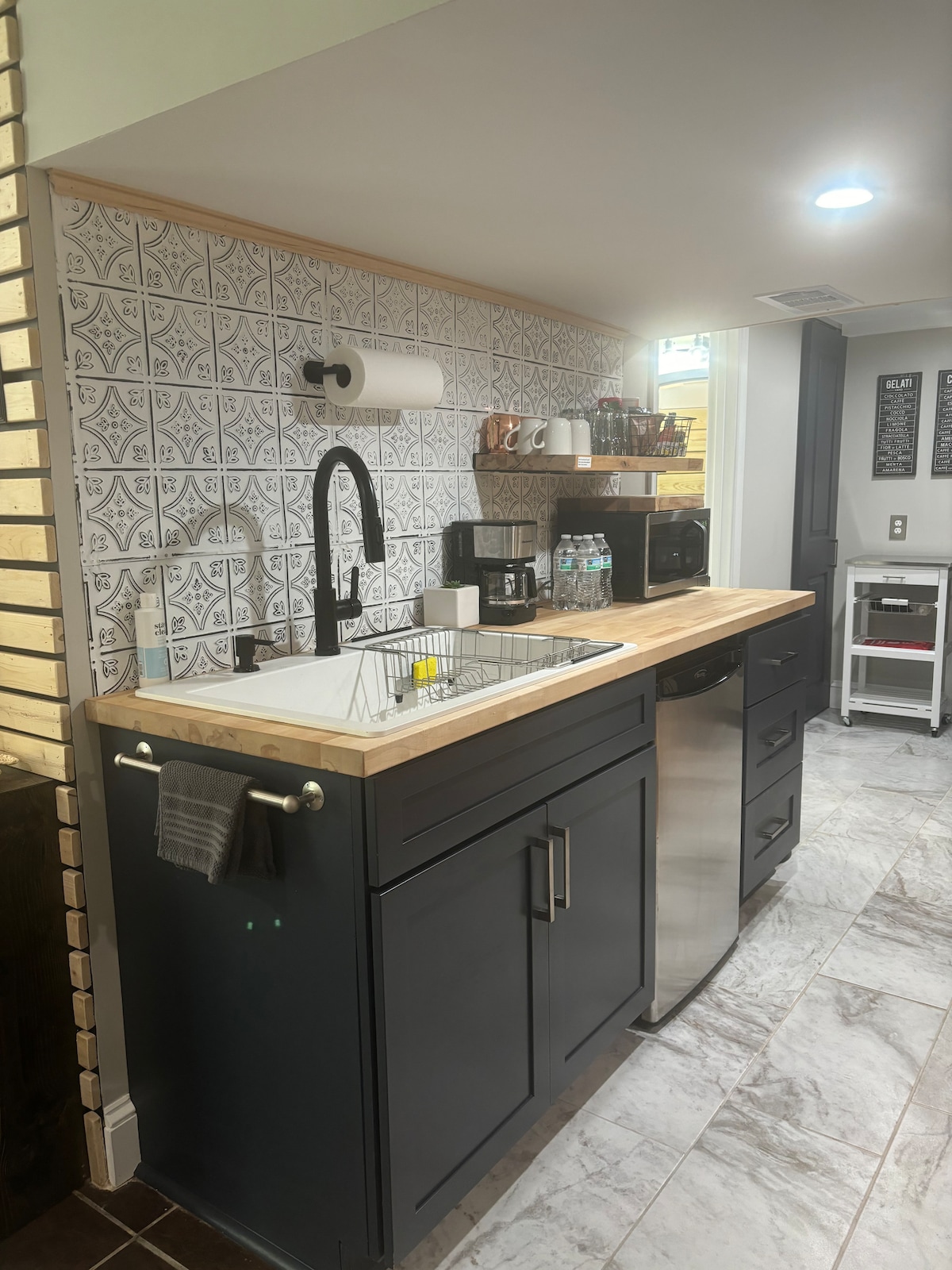 A compact kitchen area features dark cabinetry and a wooden countertop. A sink with a modern faucet is visible, along with a microwave and a water dispenser. Decorative tiles add texture to the wall, while natural light enters from an unseen source.